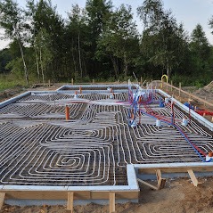 Fundament domu z rozłożonym systemem ogrzewania podłogowego, widoczne białe rurki ułożone w spiralny wzór wewnątrz drewnianego szalunku, z wystającymi rurkami instalacji wodnej i kanalizacyjnej...