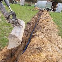 Wykonany wykop pod kabel elektryczny z ułożonym przewodem w ziemi. Widoczna łyżka koparki i otoczenie trawnika.