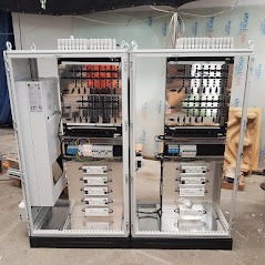 Dwie otwarte szafy elektryczne w trakcie montażu, widoczne okablowanie, listwy zaciskowe, zasilacze i inne komponenty, ściany obłożone płytami warstwowymi.