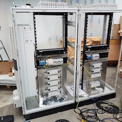 Otwarta szafa elektryczna w trakcie montażu, widoczne liczne moduły, przewody i elementy instalacyjne, wnętrze szafy oświetlone, obudowa metalowa z elementami przezroczystymi, podłoga betonowa,...