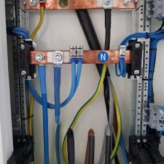 Wnętrze skrzynki rozdzielczej z widocznymi szynami miedzianymi, przewodami w izolacji niebieskiej, żółto-zielonej i czarnej, oraz elementami mocującymi na szynach DIN. Widoczny przewód neutralny...