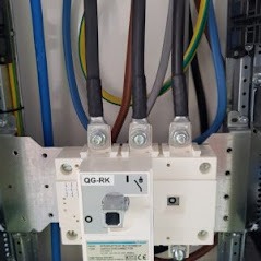 Rozdzielnica elektryczna z trzema grubymi kablami w czarnej izolacji, podłączonymi do wyłącznika głównego QG-RK. Widoczne przewody w kolorach niebieskim i zielono-żółtym. Metalowa obudowa...