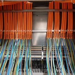 Rozdzielnica elektryczna z uporządkowanymi pomarańczowymi przewodami oznaczonymi etykietami i luźno wiszącymi niebiesko-zielonymi przewodami.