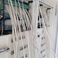 Rozdzielnica elektryczna w trakcie montażu: widoczne liczne białe przewody, częściowo podłączone do listew zaciskowych, pozostałe swobodnie zwisają. Ściana w tle w kolorze białym.