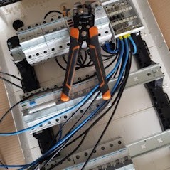 Otwarta skrzynka elektryczna z zamontowanymi bezpiecznikami i podłączonymi przewodami. Na wierzchu leżą szczypce do zdejmowania izolacji z pomarańczowymi uchwytami.