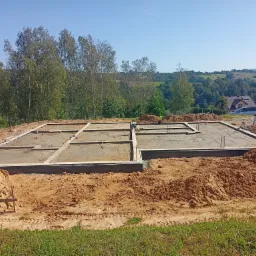 Wylany fundament pod budowę domu jednorodzinnego, widoczne zbrojenia i szalunki, w tle krajobraz z drzewami i zabudowaniami.