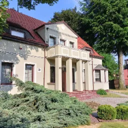 Elewacja domu jednorodzinnego z beżową fasadą, czerwonym dachem i balkonem wspartym na kolumnach, z numerem domu '86' nad wejściem, otoczona zielenią i kostką brukową.