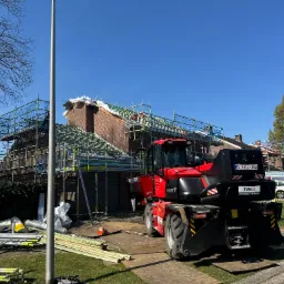 Ceglany dom w trakcie prac dekarskich, otoczony rusztowaniem, z widocznym czerwonym dźwigiem teleskopowym i materiałami budowlanymi na trawniku.