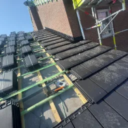 Układanie czarnej dachówki na drewnianej konstrukcji dachu, widoczne narzędzia dekarskie i rusztowanie przy ceglanym budynku w słoneczny dzień.