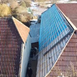 BART-DACH - Widok z góry na budynek z częściowo zdjętym dachem i widoczną konstrukcją więźby dachowej pokrytą niebieską folią, sąsiednie budynki z dachami z czerwonej dachówki karpiówki.