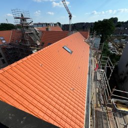 BART-DACH - Pomarańczowa dachówka ceramiczna na dachu budynku z oknem dachowym, widok z góry na rusztowania i krajobraz miejski z żurawiami budowlanymi w tle.