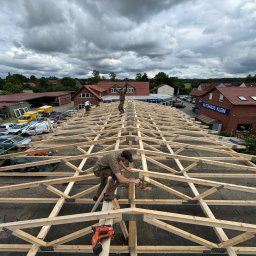 BART-DACH - Montaż drewnianej więźby dachowej z użyciem gwoździarki pneumatycznej, widok z góry na konstrukcję i dwóch dekarzy pracujących na dachu, w tle zabudowa miejska i pochmurne niebo.