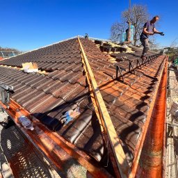 BART-DACH - Pracownik na dachu układa brązowe dachówki, widoczne rusztowanie, zabezpieczenia i narzędzia, błękitne niebo w tle.