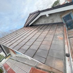 BART-DACH - Ukończony fragment dachu pokryty ciemną dachówką, widoczny z rusztowania, z elementami obróbek blacharskich i fragmentem okna dachowego zabezpieczonego folią.