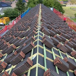 BART-DACH - Widok z góry na spadzisty dach w trakcie prac, z licznymi stosami ciemnobrązowych dachówek na drewnianych łatach i czarnej membranie, w tle domy i zieleń.