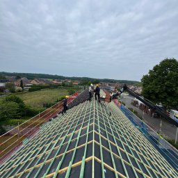BART-DACH - Trzech dekarzy w żółtych kaskach pracuje na dachu, układając ciemne dachówki. Widoczna konstrukcja z zielonych łat i czarnej membrany. Żuraw podaje materiały.