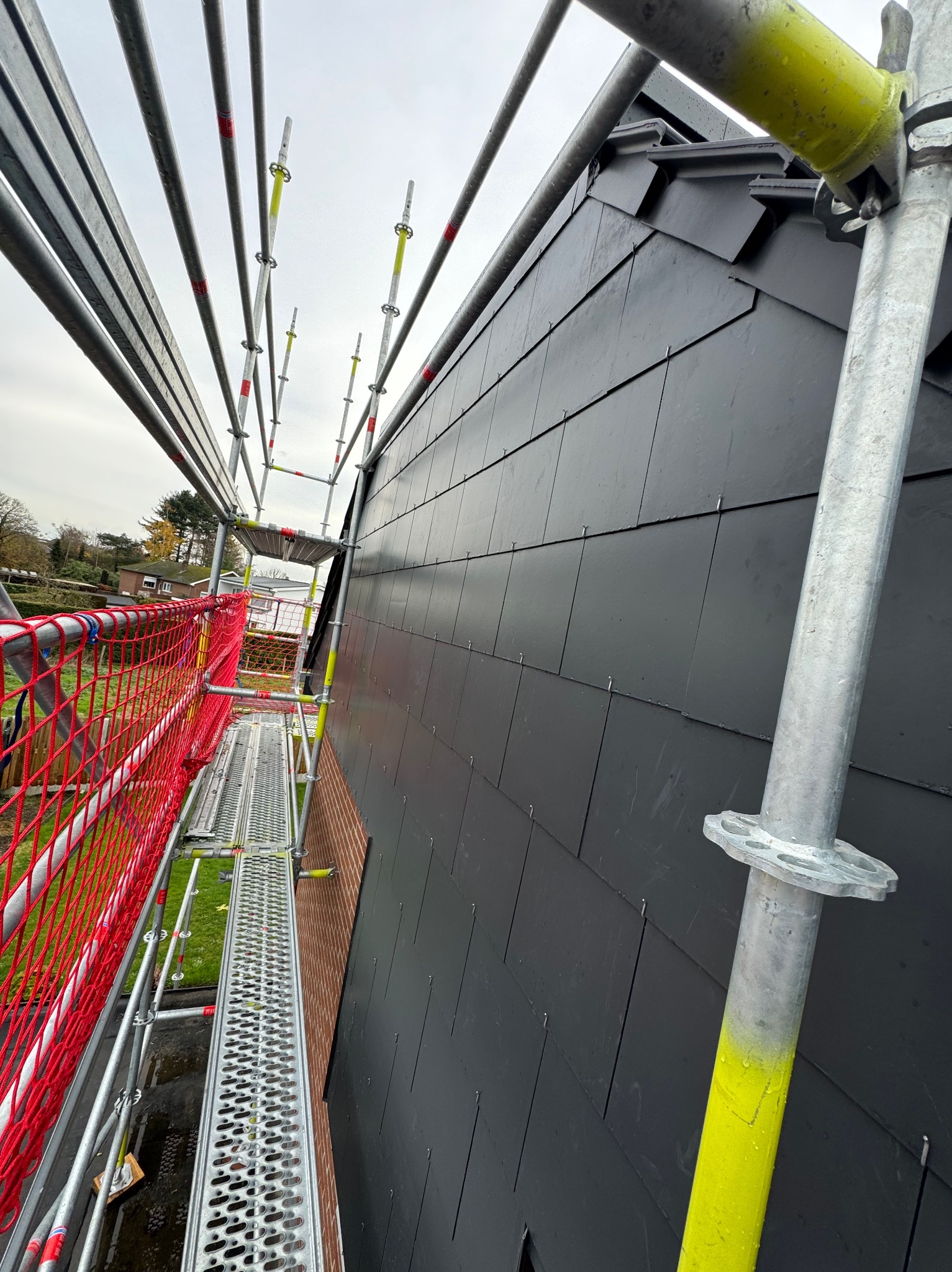 Ciemnoszary dach z łupka w trakcie montażu, widok z rusztowania z pomalowanymi na żółto elementami, siatka zabezpieczająca w kolorze czerwonym.