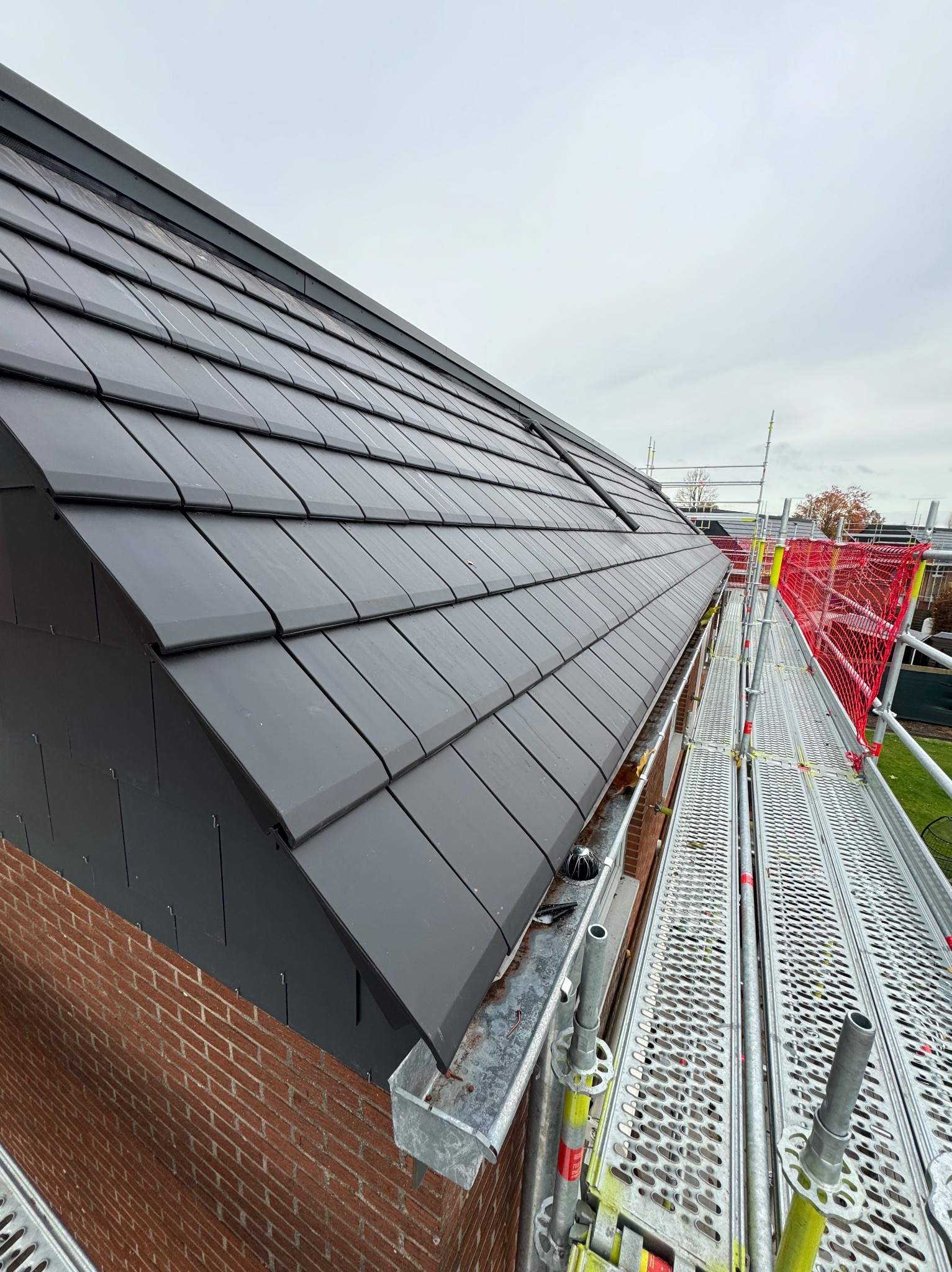 Ciemnoszara dachówka ceramiczna ułożona na dachu z ceglaną ścianą boczną, widok z rusztowania z zabezpieczeniem siatkowym.
