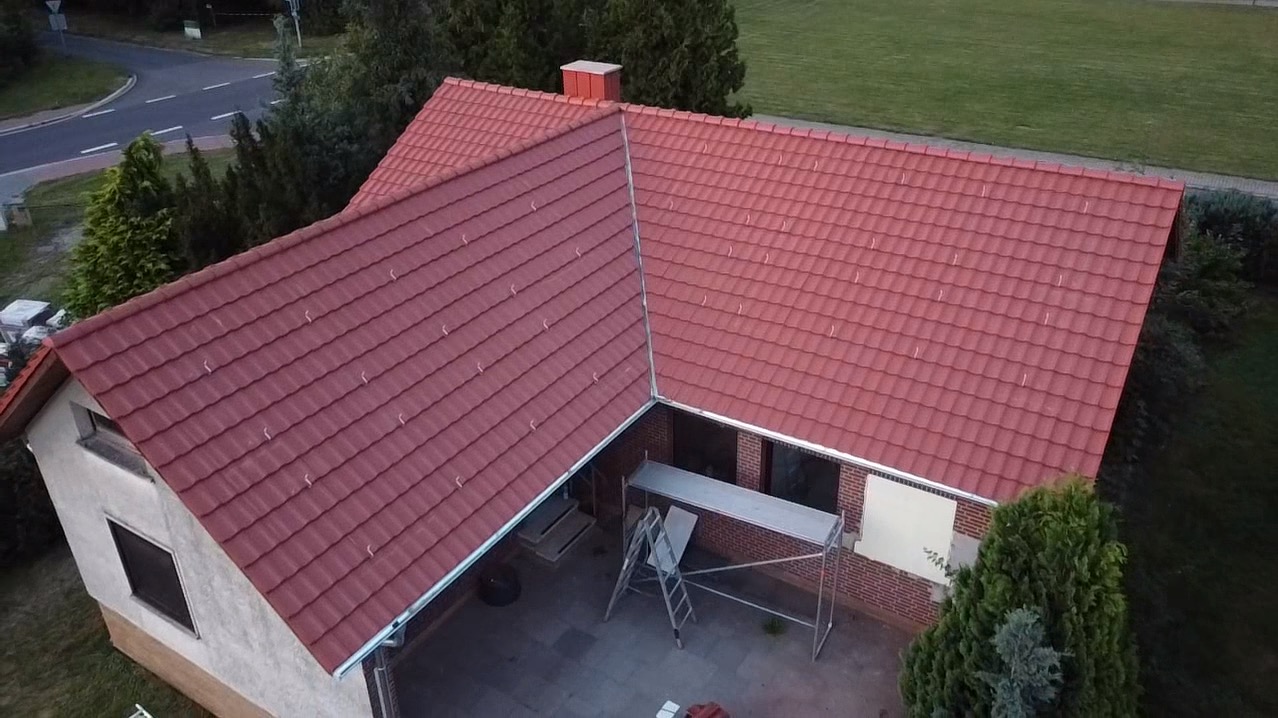 Widok z góry na dom z nowym, czerwonym dachem z dachówki ceramicznej; widoczne rusztowanie i drabina przy ścianie budynku.