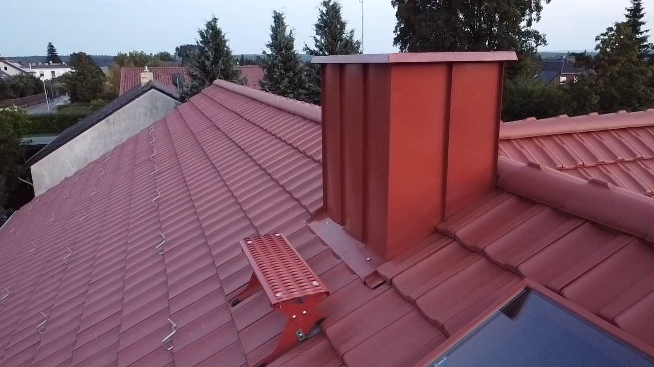 Nowy, czerwony dach z dachówki ceramicznej z kominem w tym samym kolorze, zabezpieczeniem przeciwpoślizgowym i oknem dachowym, widok z góry.