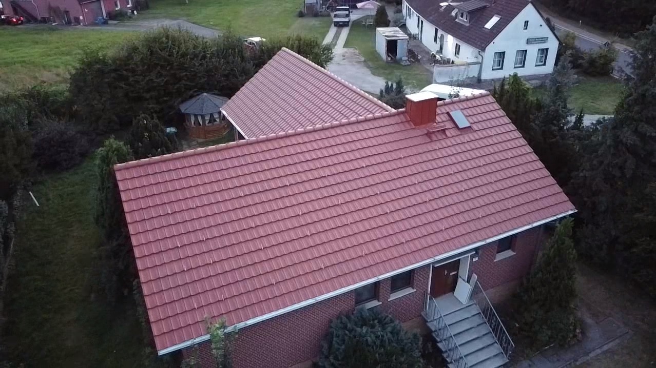 Widok z góry na dom z nowym, czerwonym dachem z dachówki ceramicznej, z kominem i oknem dachowym, otoczony zielenią i zabudowaniami gospodarczymi.