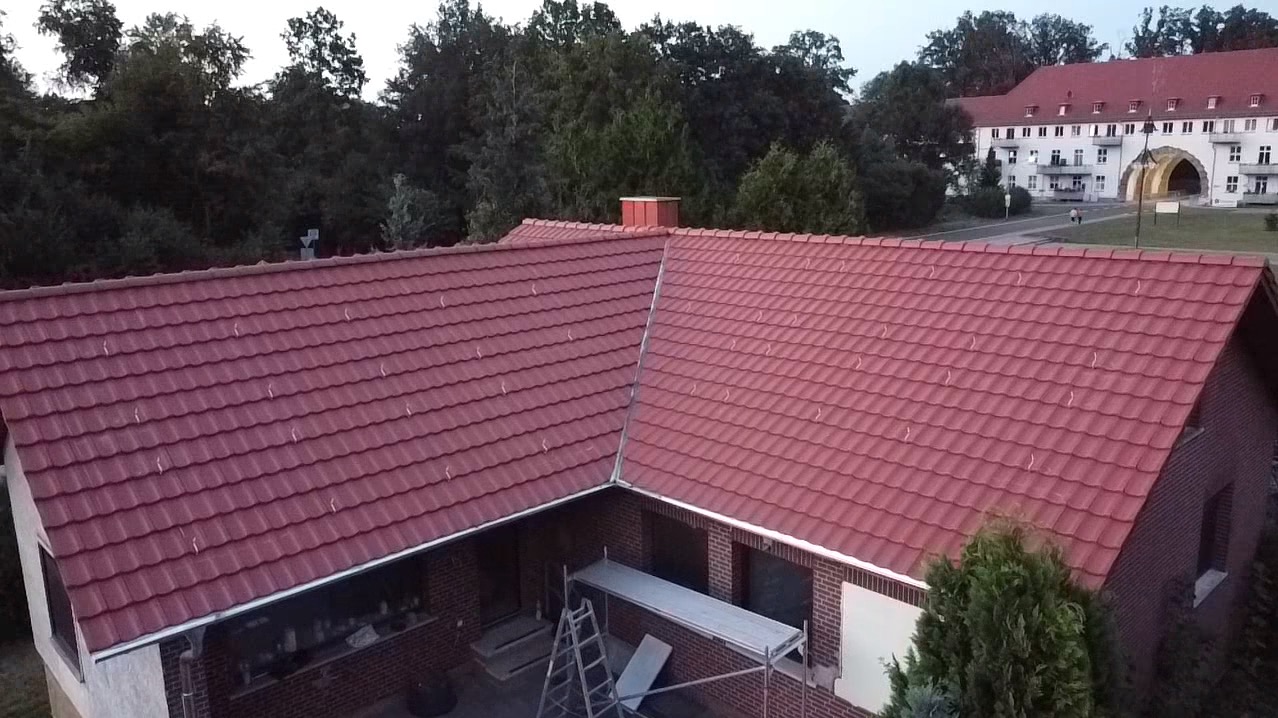 Czerwony dach z dachówki ceramicznej, widok z góry na budynek z cegły z rusztowaniem i drabiną, w tle budynek z łukowym wejściem i drzewa.