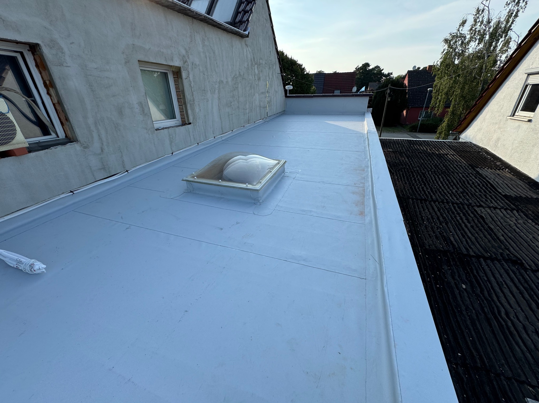 Szary płaski dach z świetlikiem kopułowym, widok z góry na tle sąsiednich budynków i drzew