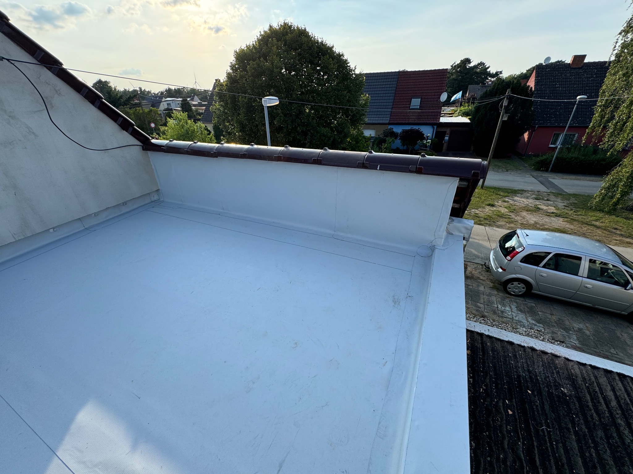 Widok z góry na płaski, biały dach pokryty papą termozgrzewalną, z widocznymi brązowymi elementami wykończeniowymi i fragmentem dachu sąsiedniego budynku oraz samochodem osobowym w tle.