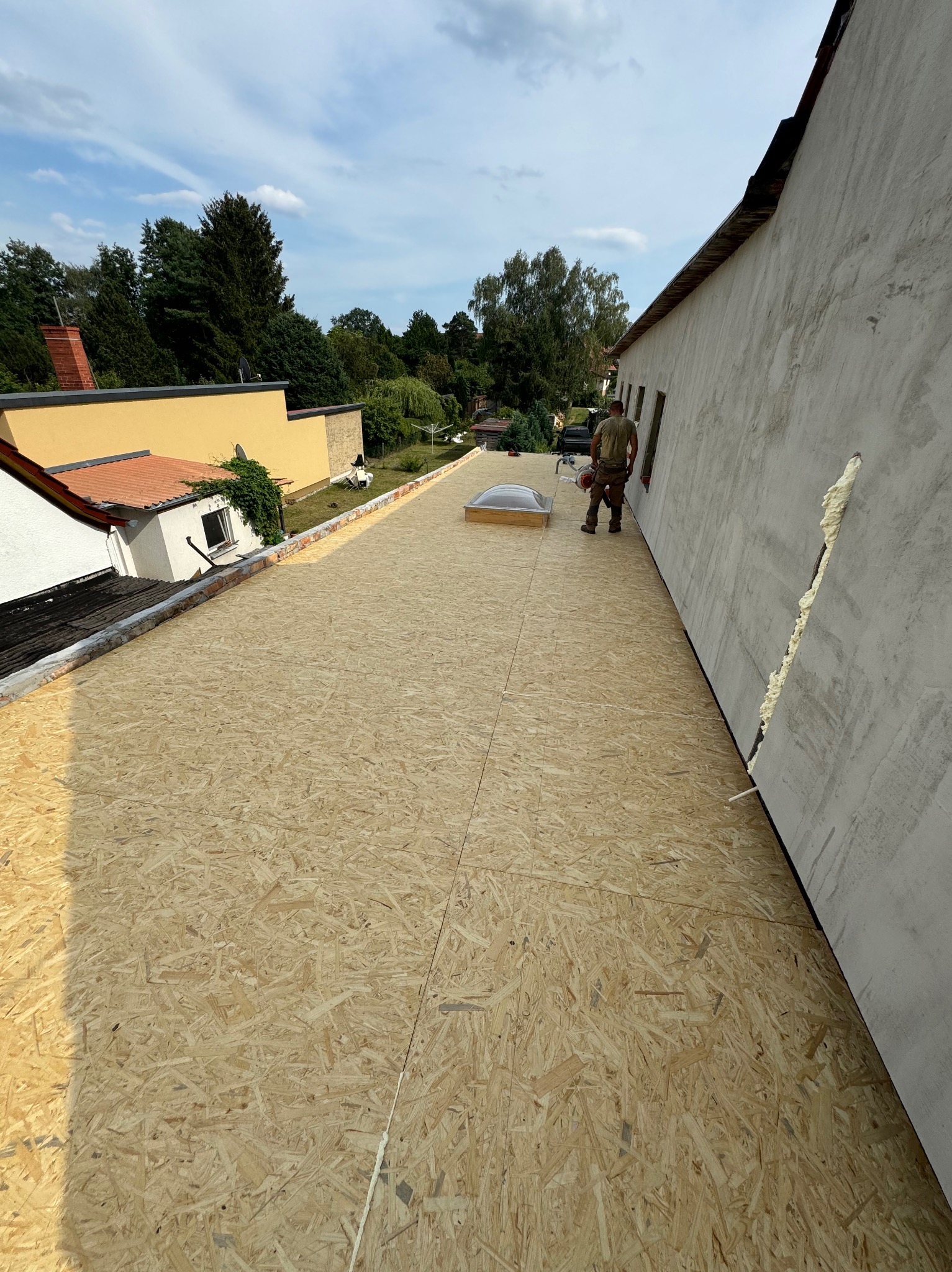 Płyty OSB ułożone na dachu z widocznym świetlikiem dachowym i pracownikiem w tle, widok z góry na przygotowany dach przed dalszymi pracami dekarskimi.