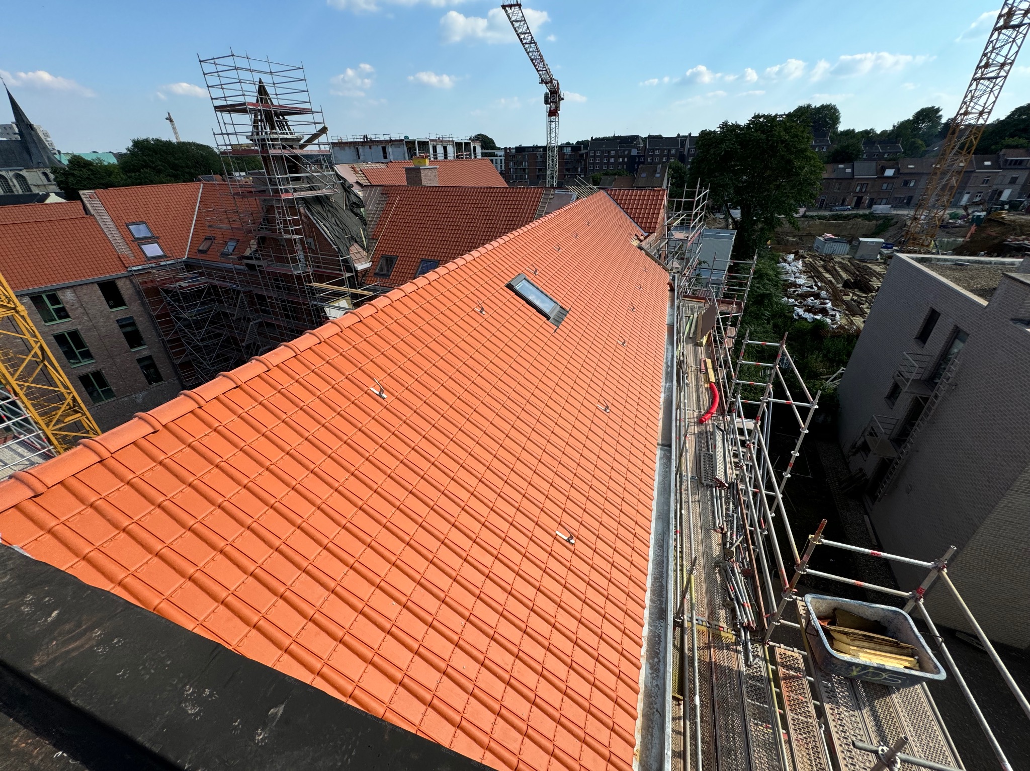 Pomarańczowa dachówka ceramiczna na dachu budynku z oknem dachowym, widok z góry na rusztowania i krajobraz miejski z żurawiami budowlanymi w tle.