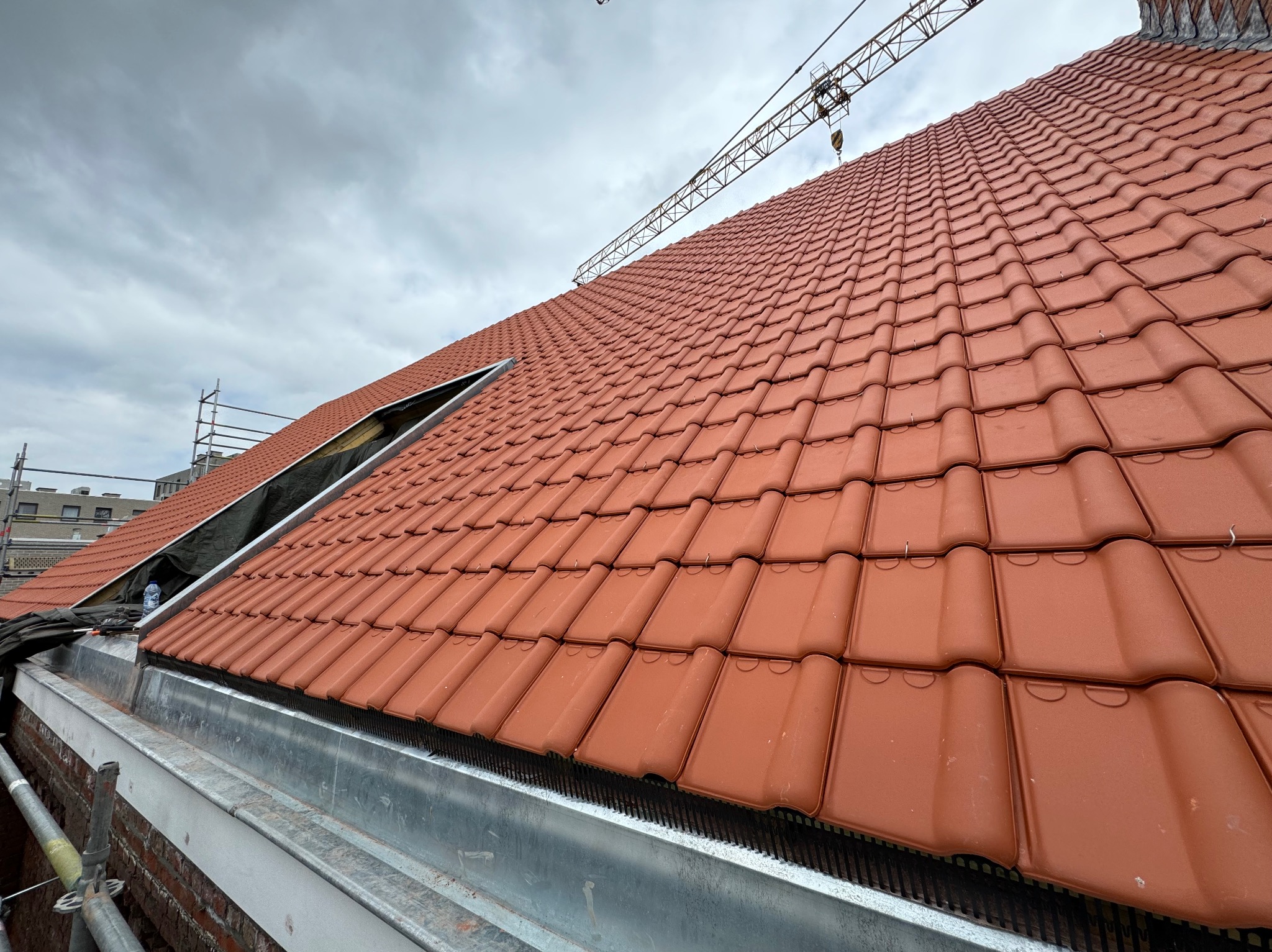 Nowe, czerwone dachówki ceramiczne ułożone na dachu, z widocznym fragmentem rusztowania i żurawiem w tle, pod pochmurnym niebem.