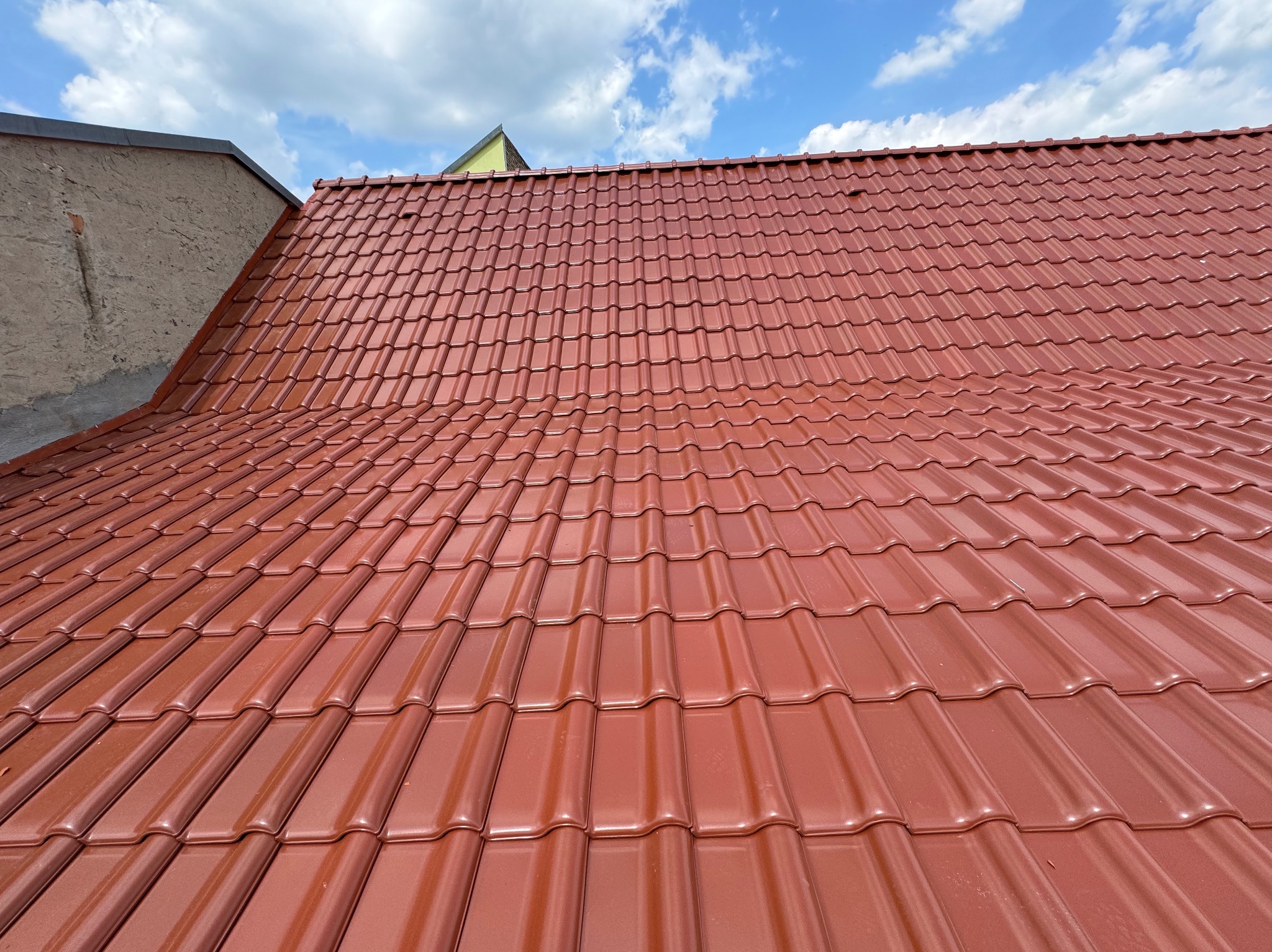 Czerwona dachówka ceramiczna na ukośnym dachu w słoneczny dzień, widok z dołu, fragment sąsiedniego budynku w tle.