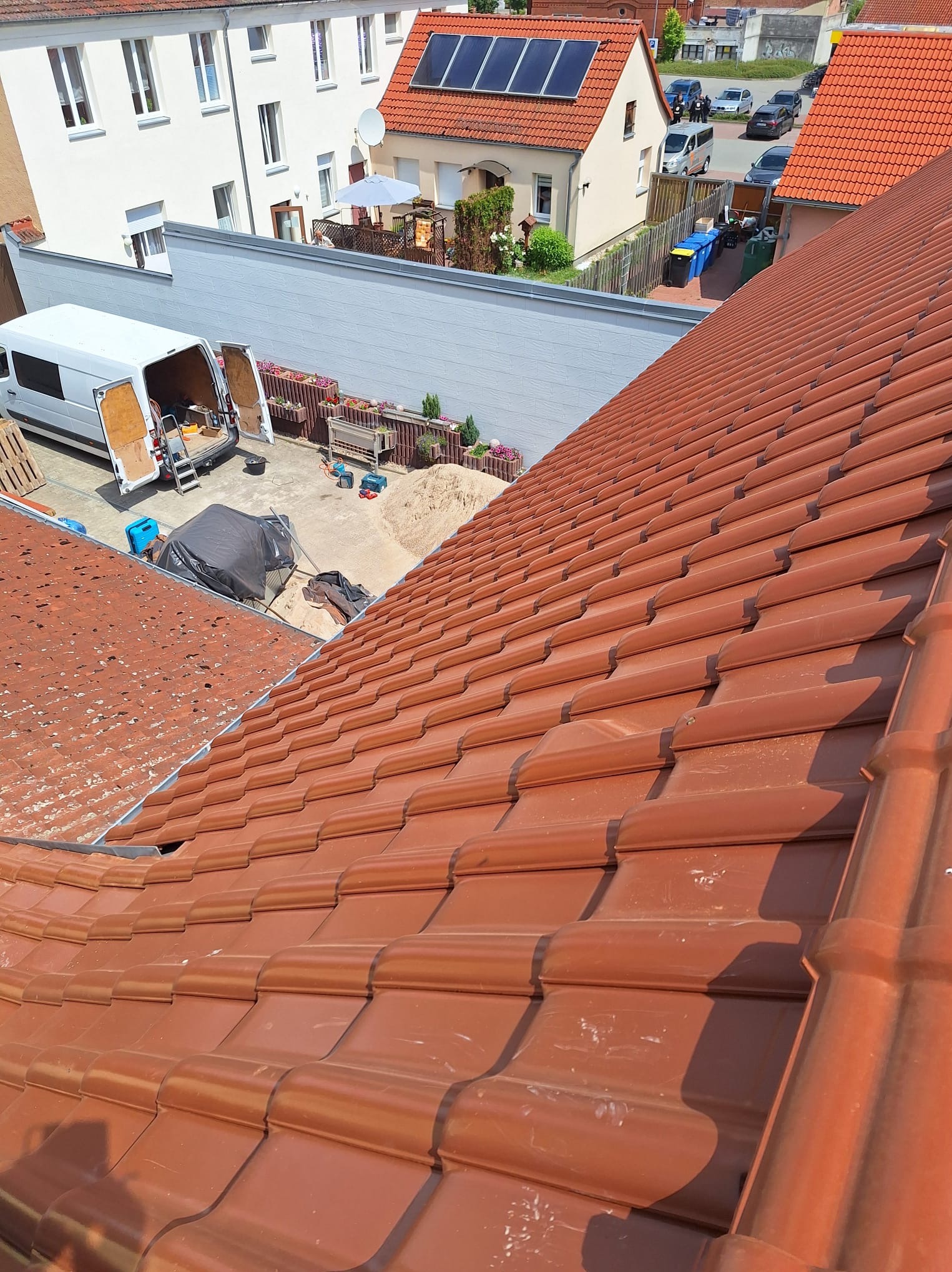 Ukośny widok na dach z czerwonej dachówki ceramicznej, w tle biały van z otwartymi drzwiami, sprzęt budowlany i domy mieszkalne.