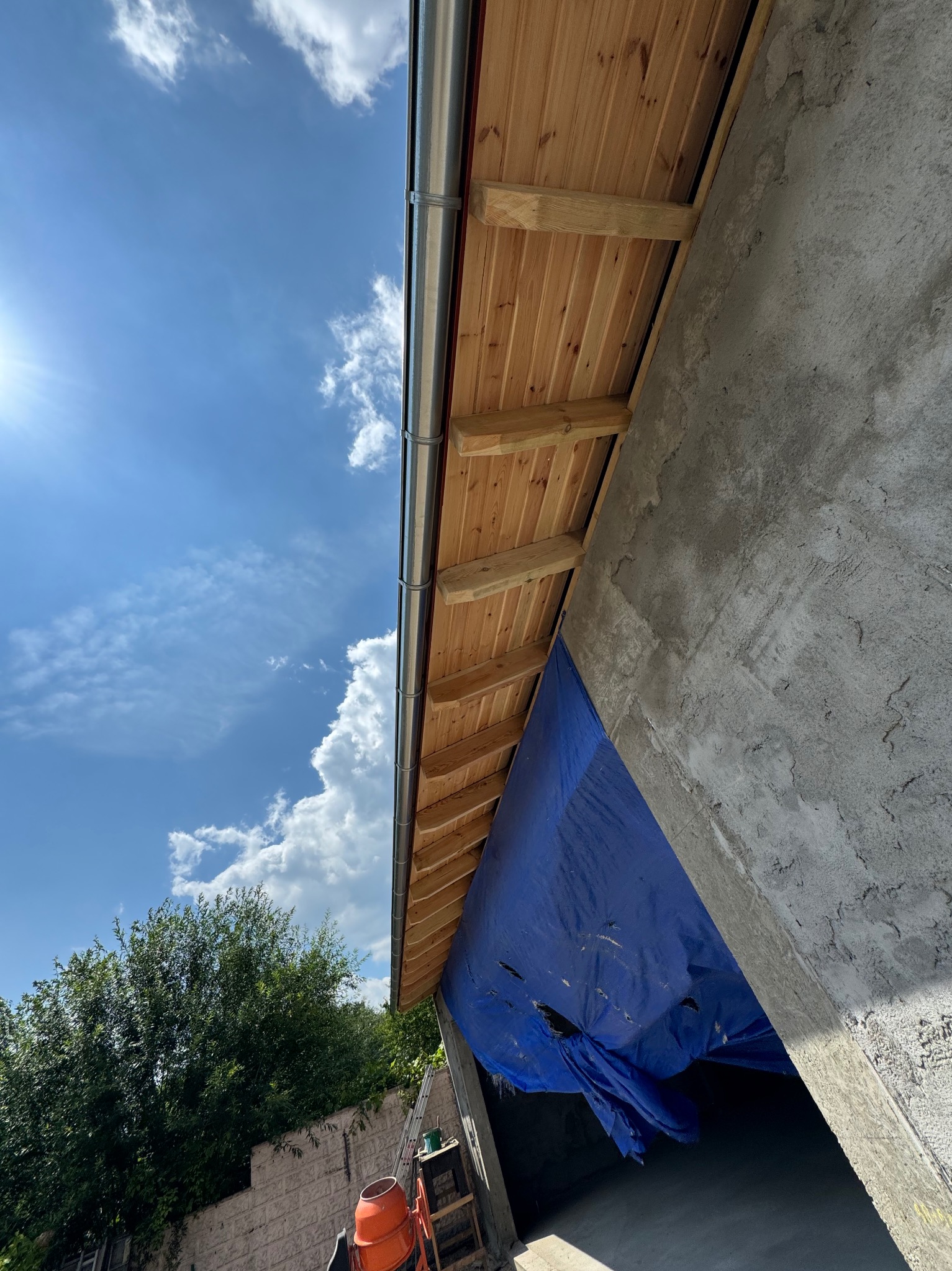 Drewniana podbitka dachowa z zamontowaną metalową rynną, widok od dołu na tle błękitnego nieba z chmurami, fragment surowego muru z plandeką zabezpieczającą.