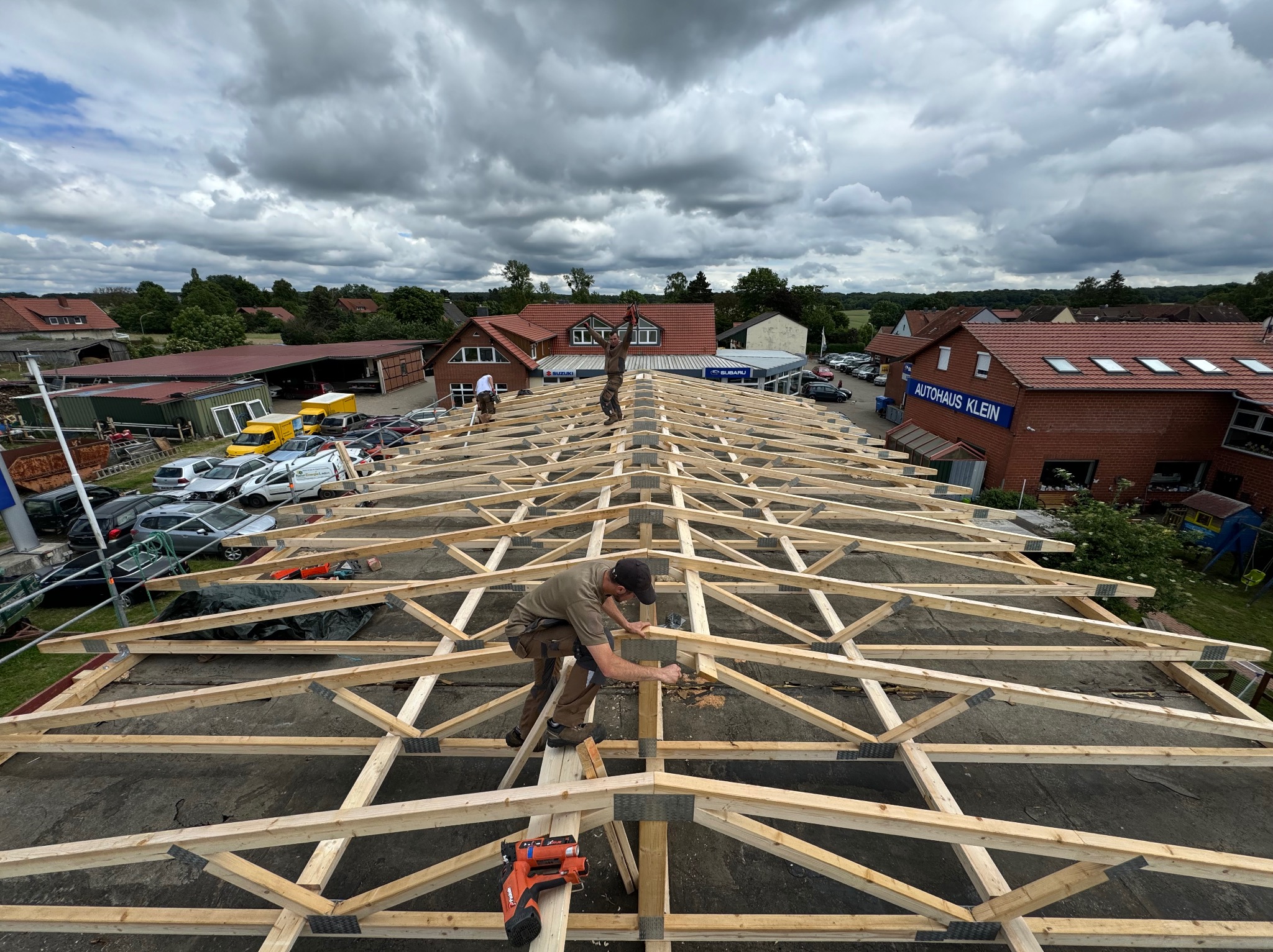 Montaż drewnianej więźby dachowej z użyciem gwoździarki pneumatycznej, widok z góry na konstrukcję i dwóch dekarzy pracujących na dachu, w tle zabudowa miejska i pochmurne niebo.