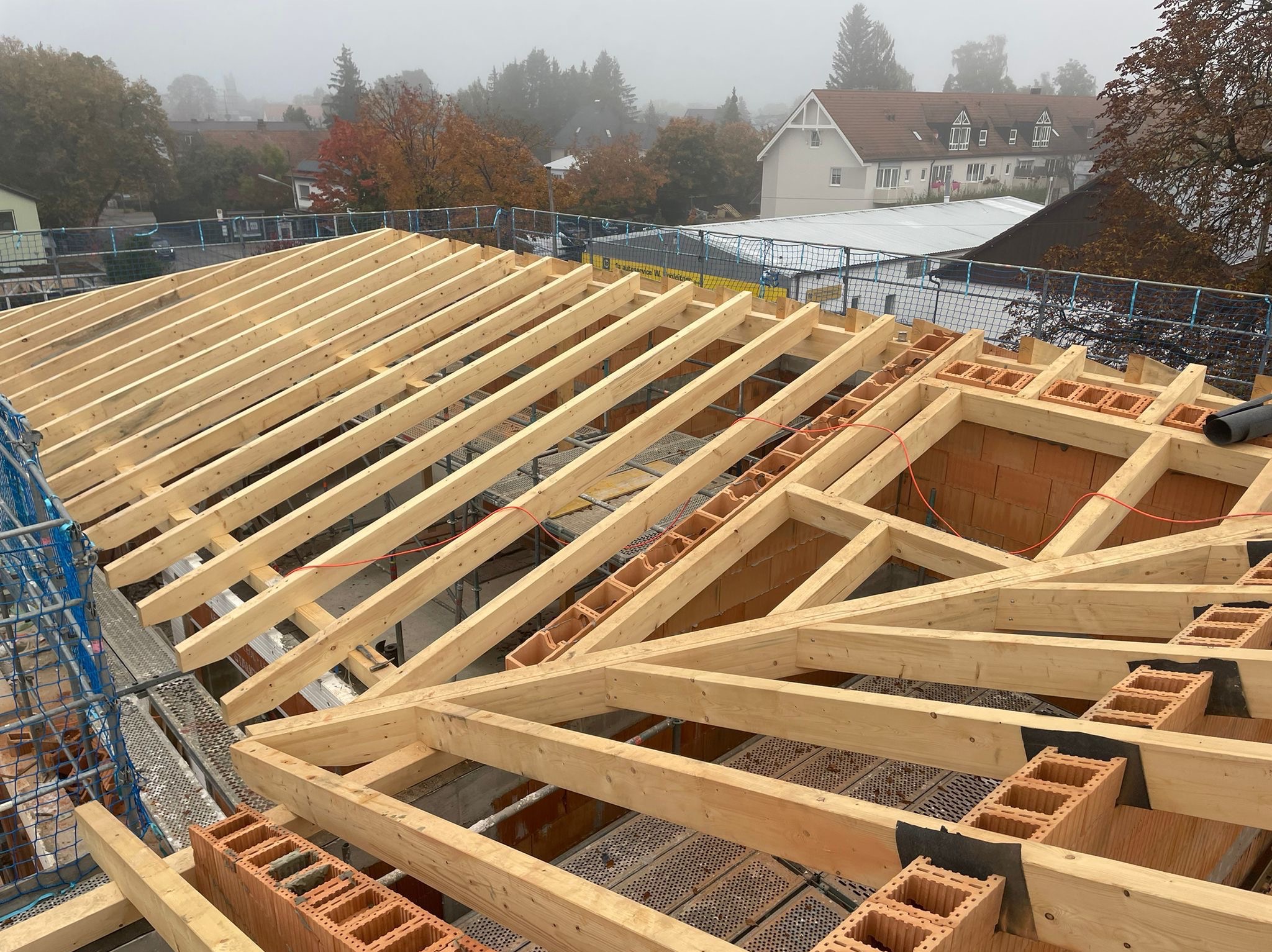 Drewniana konstrukcja więźby dachowej w trakcie montażu, widoczne cegły dziurawki i elementy zabezpieczeń na tle zamglonego krajobrazu miejskiego.