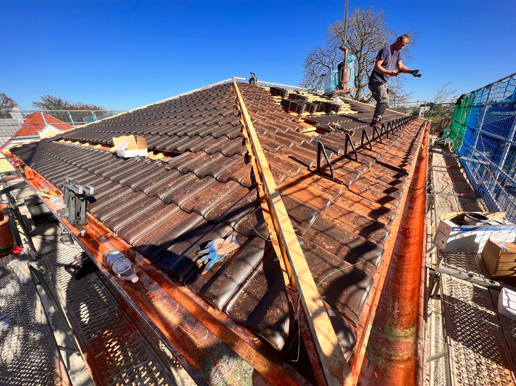 Pracownik na dachu układa brązowe dachówki, widoczne rusztowanie, zabezpieczenia i narzędzia, błękitne niebo w tle.