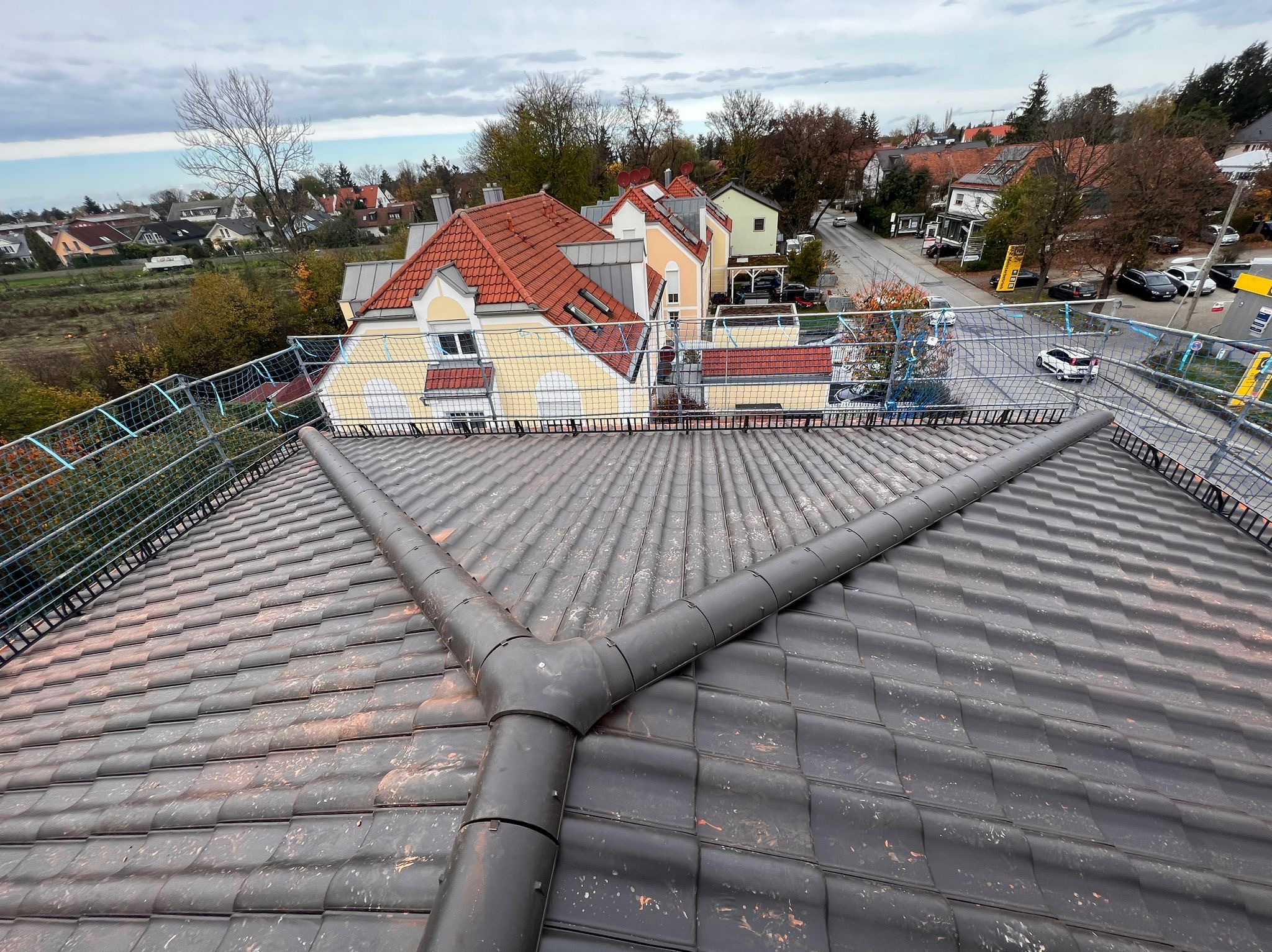 Widok z góry na ciemną dachówkę ceramiczną z trójnikiem odprowadzającym wodę, zabezpieczoną siatką ochronną, w tle zabudowa jednorodzinna i pochmurne niebo.
