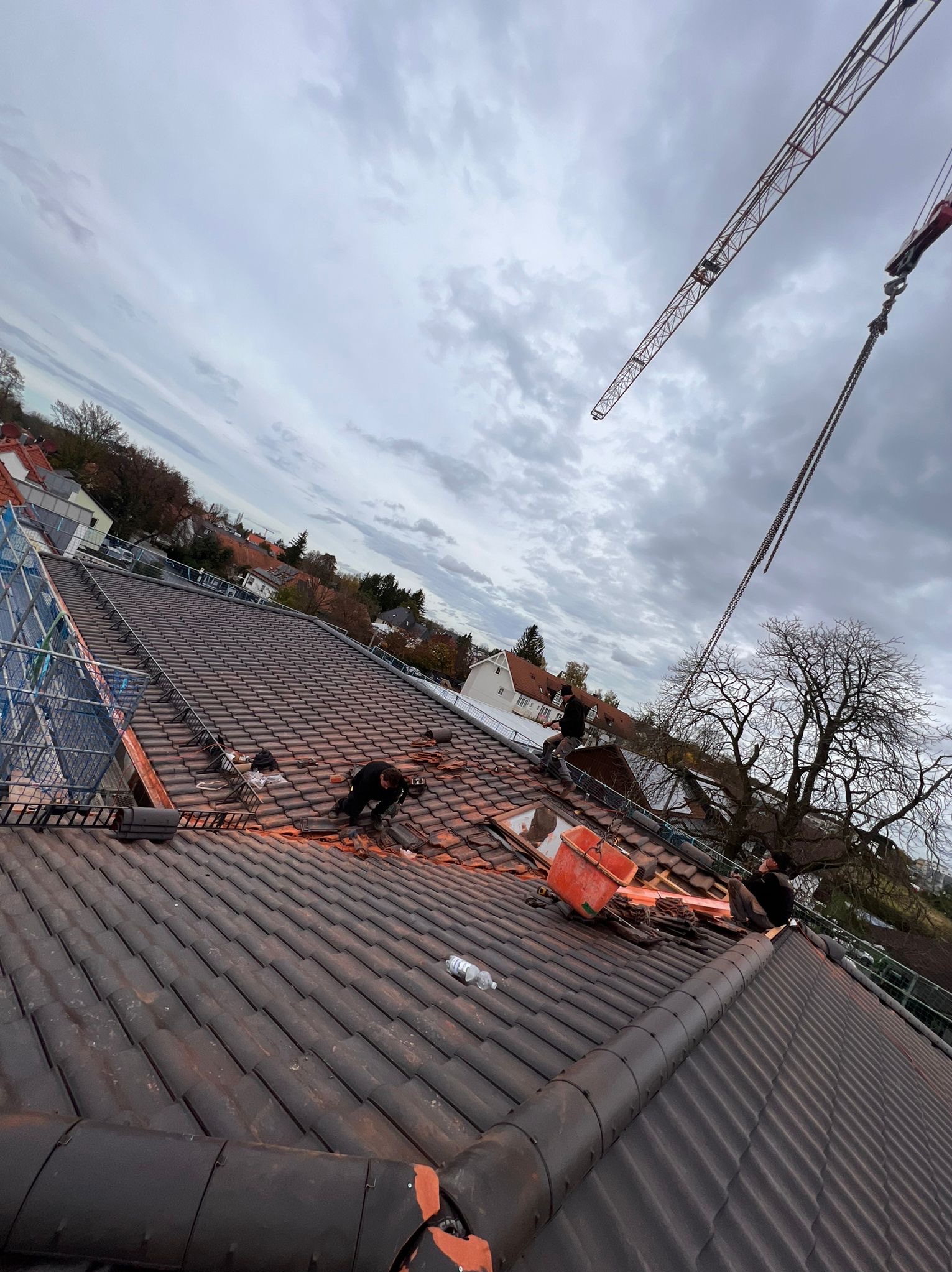 Ekipa dekarzy pracuje na dachu z ciemnej dachówki ceramicznej, widoczny dźwig budowlany i drabina, pochmurne niebo w tle.