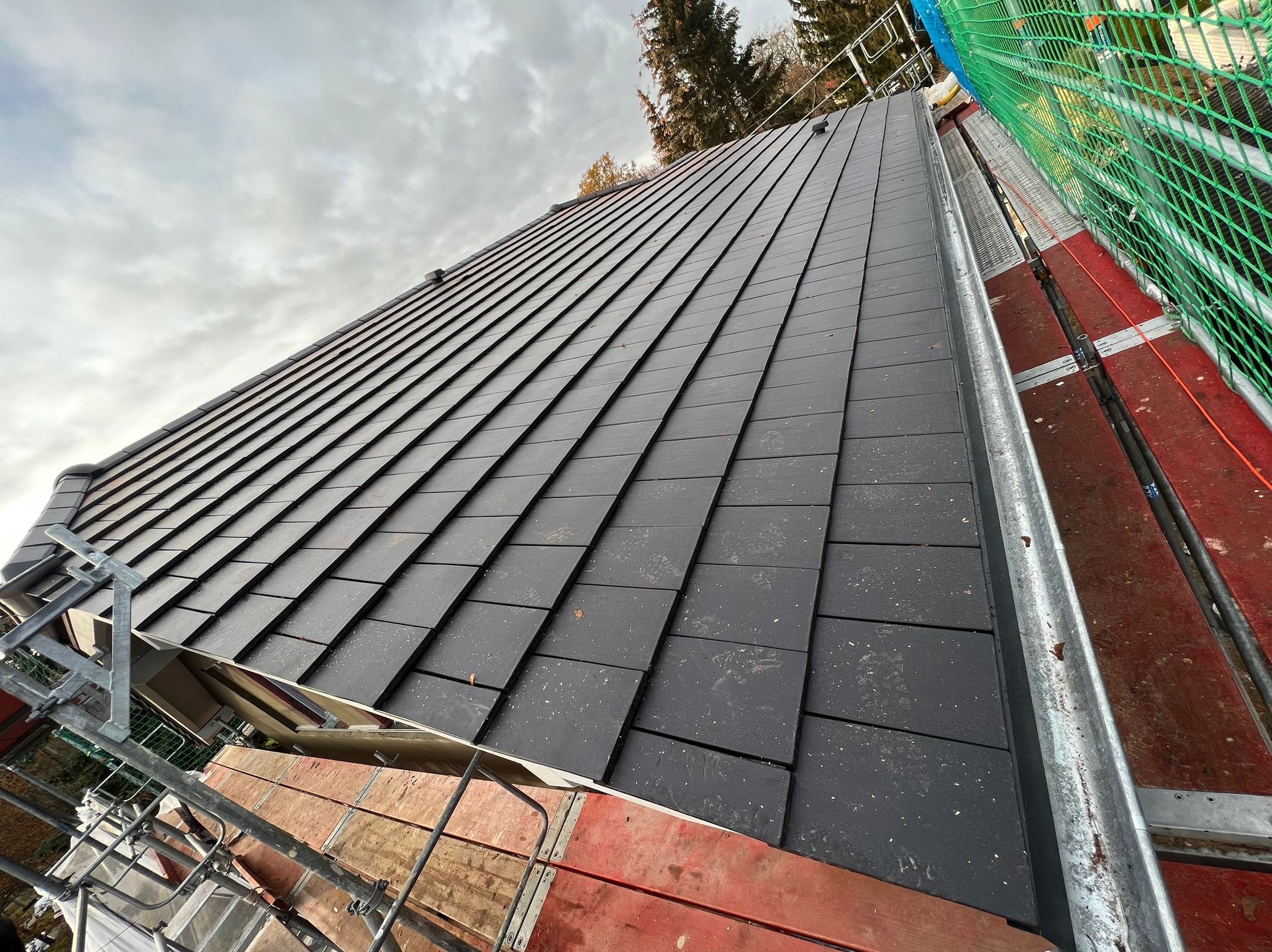 Nowy, ciemnoszary dach z dachówki ceramicznej widziany z rusztowania, z elementami zabezpieczeń i siatki ochronnej w tle pochmurnego nieba.