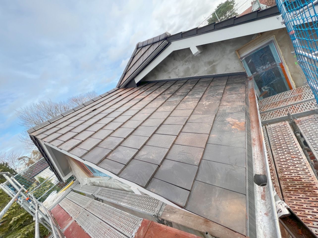 Ukończony fragment dachu pokryty ciemną dachówką, widoczny z rusztowania, z elementami obróbek blacharskich i fragmentem okna dachowego zabezpieczonego folią.