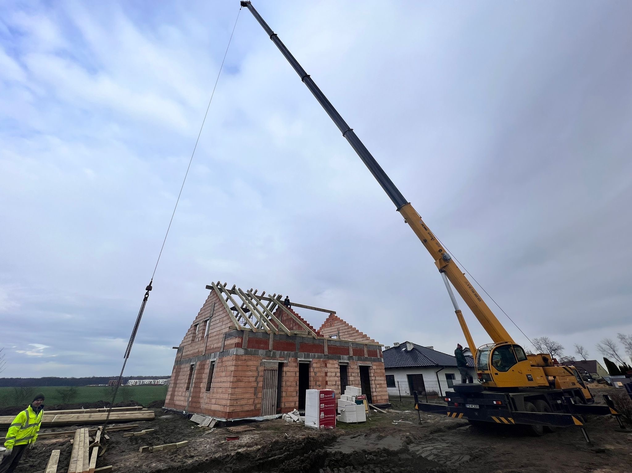 Budowa domu jednorodzinnego w trakcie montażu więźby dachowej przy użyciu dźwigu, widoczny mur z czerwonej cegły, pracownik w odblaskowej kamizelce oraz materiały budowlane na placu budowy.