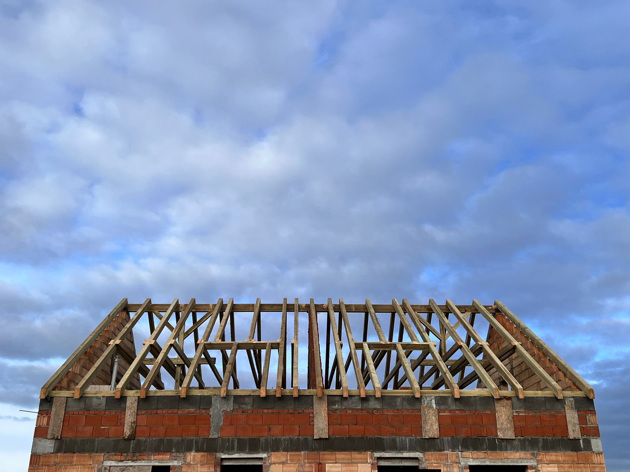 Konstrukcja drewnianej więźby dachowej na murowanym budynku z czerwonej cegły, na tle pochmurnego, błękitnego nieba.