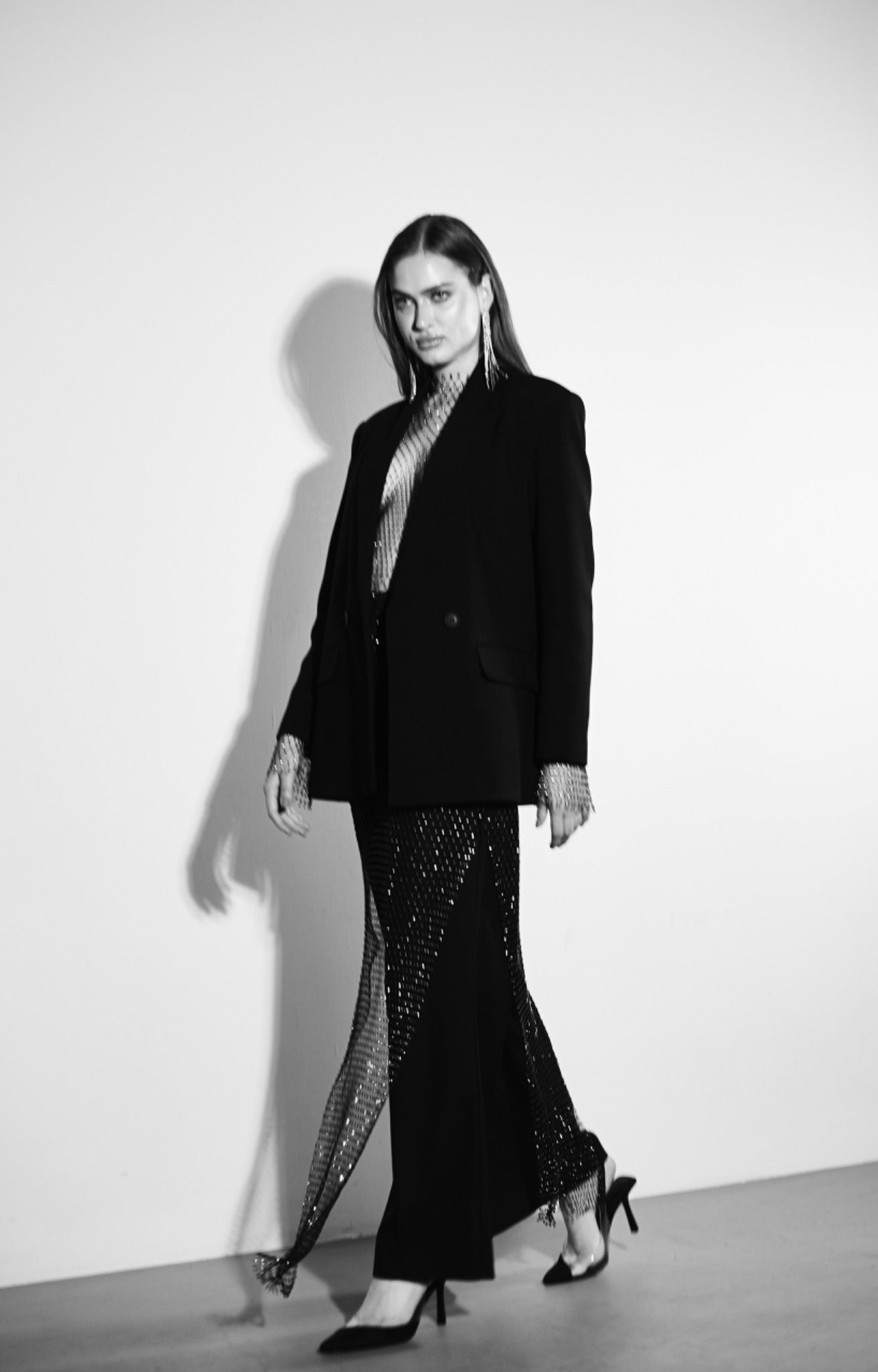 Czarno-białe zdjęcie modelki w czarnym oversize'owym żakiecie i długiej, błyszczącej spódnicy z rozcięciem, pozującej na jasnym tle. Długie kolczyki i ciemne włosy dopełniają elegancki wygląd.
