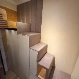 Schody z obiciem tapicerskim prowadzące na antresolę, zintegrowane z szafkami, oświetleniem LED i tapicerowanym panelem ściennym w kolorze beżowym.