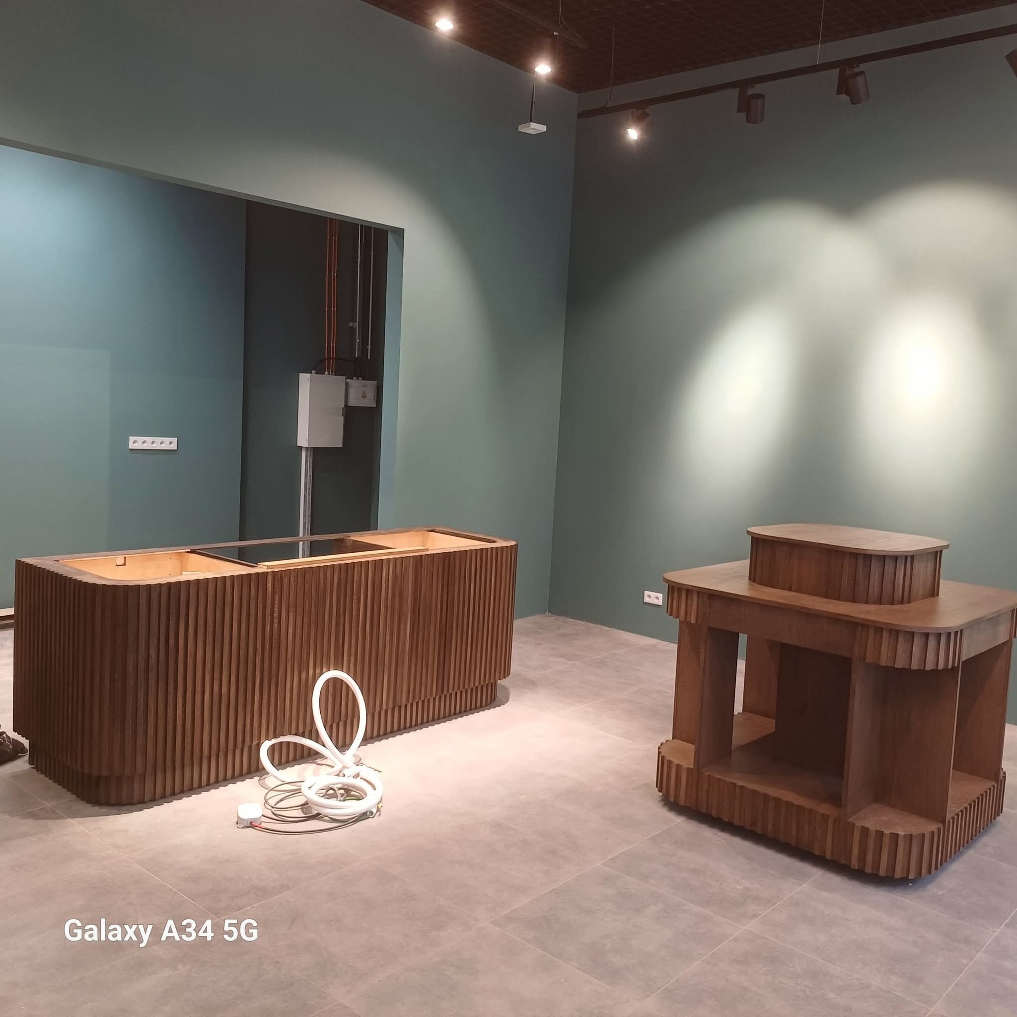 Brązowe, ryflowane meble w surowym wnętrzu z zielonymi ścianami i szarą podłogą. Na podłodze leżą białe kable. Widoczne oświetlenie sufitowe.