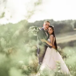 Para młoda w objęciach na tle zieleni, panna młoda w koronkowej sukni z długimi włosami, pan młody w szarej kraciastej marynarce i muszce, bukiet w rękach, delikatne światło.