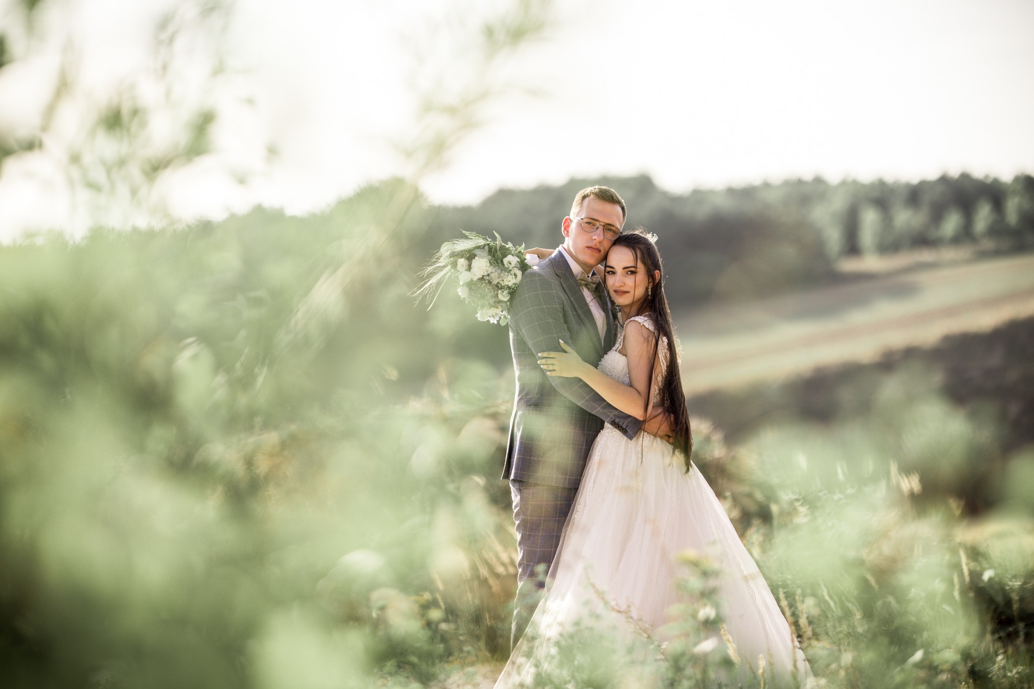 Para młoda w objęciach na tle zieleni, panna młoda w koronkowej sukni z długimi włosami, pan młody w szarej kraciastej marynarce i muszce, bukiet w rękach, delikatne światło.