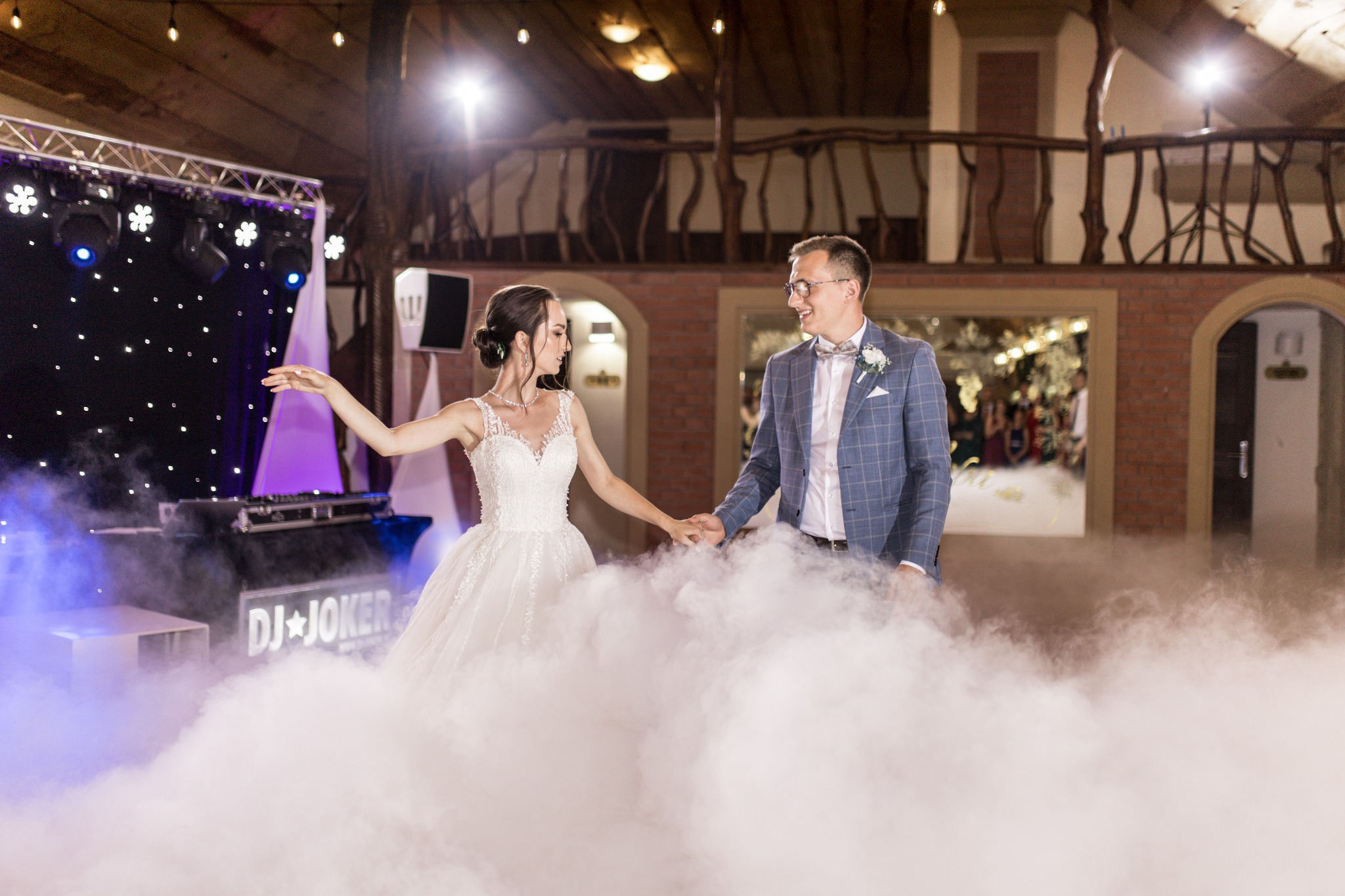 Para młoda trzymająca się za ręce w tańcu, otoczona sztuczną mgłą na tle sceny z oświetleniem DJ'a i rustykalnego balkonu.