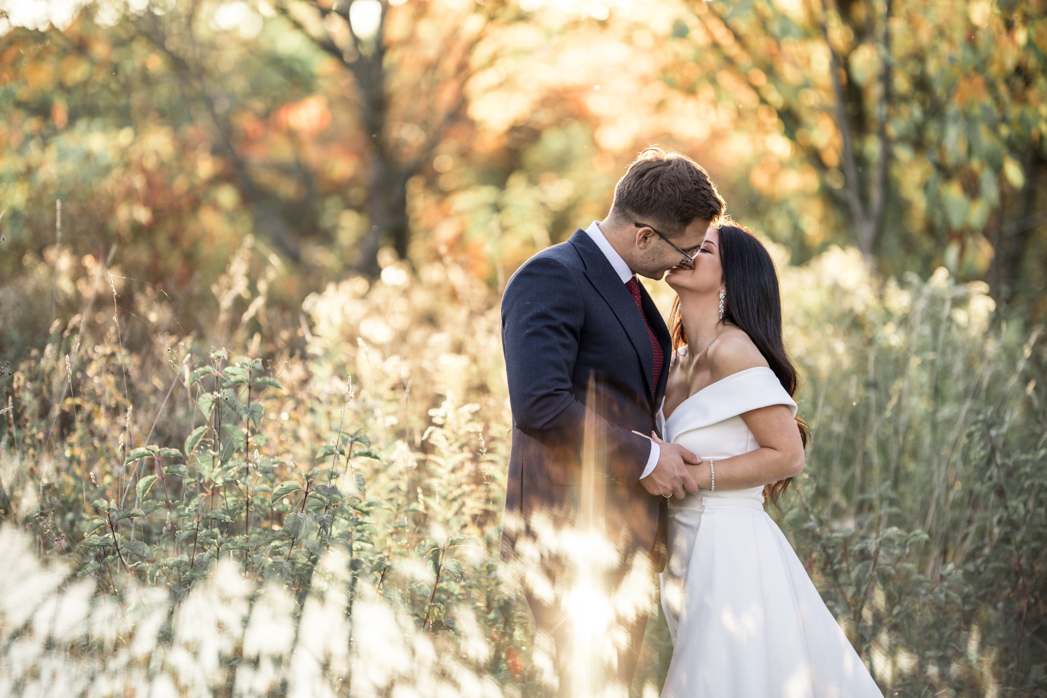 Para młoda całuje się na tle jesiennego krajobrazu, mężczyzna w granatowym garniturze i okularach, kobieta w białej sukni z odkrytymi ramionami.